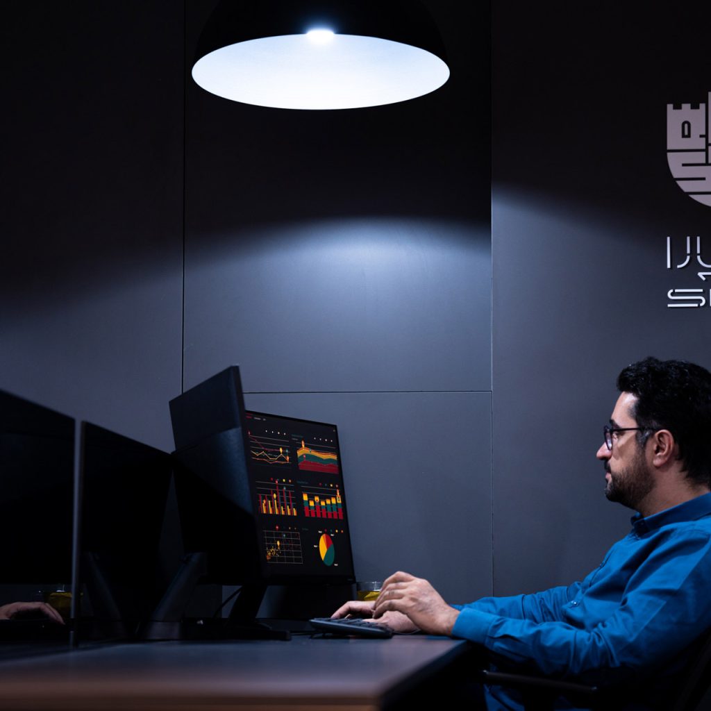 کارمند مرد در حال بررسی داده‌ها روی مانیتور در اتاق تاریک با نور متمرکز