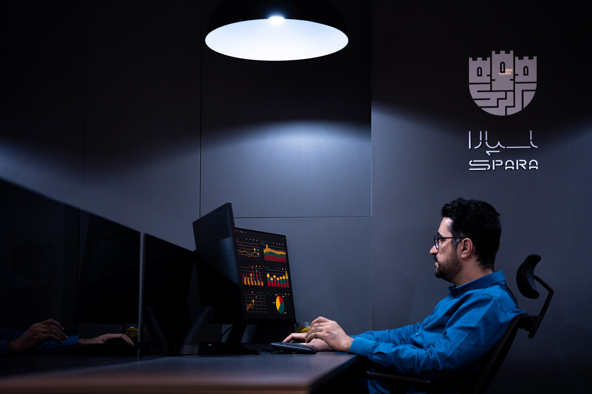 کارمند مرد در حال بررسی داده‌ها روی مانیتور در اتاق تاریک با نور متمرکز